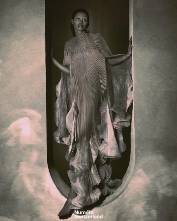 Fashion editorial shot of a Black female model posing in a pleated gown within a curved architectural frame, in a dramatic, grey-toned indoor setting.
