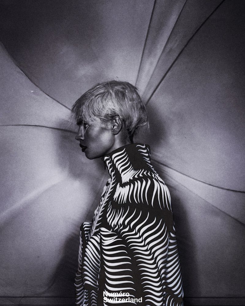 Black and white fashion editorial portrait of a woman with short hair wearing a patterned high-collar top in front of a radial backdrop.