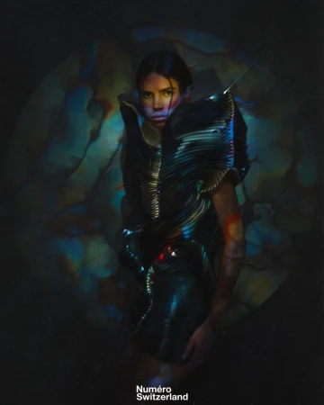 Fashion editorial shot of a woman in a futuristic black sculptural dress against a textured, colorful backdrop with dramatic lighting.