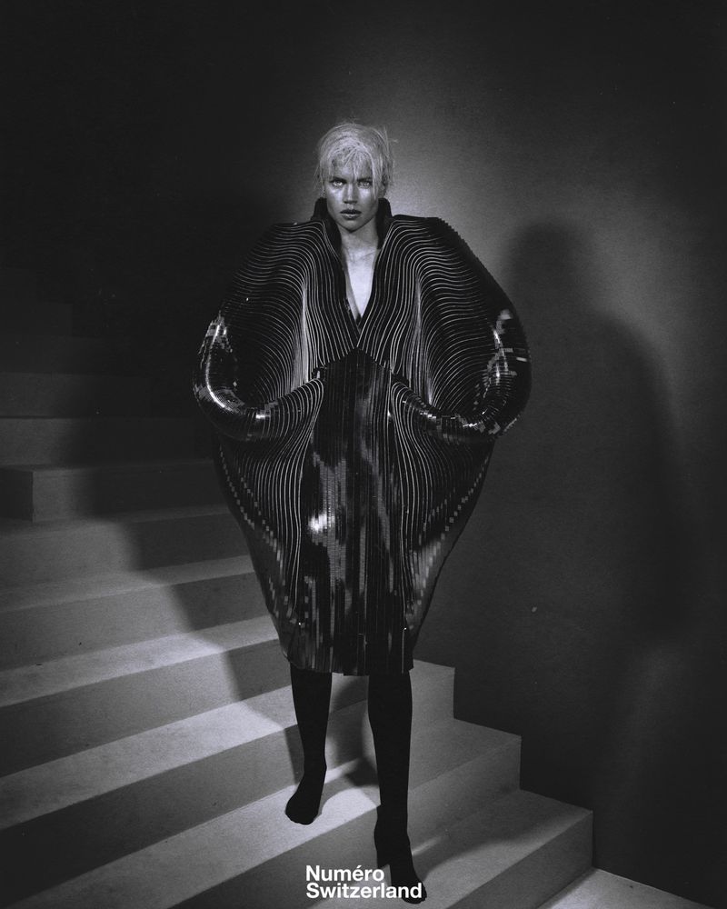 Editorial fashion shot of a model on stairs in an avant-garde dress with exaggerated sleeves and black boots, posing for the camera.