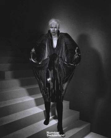 Editorial fashion shot of a model on stairs in an avant-garde dress with exaggerated sleeves and black boots, posing for the camera.