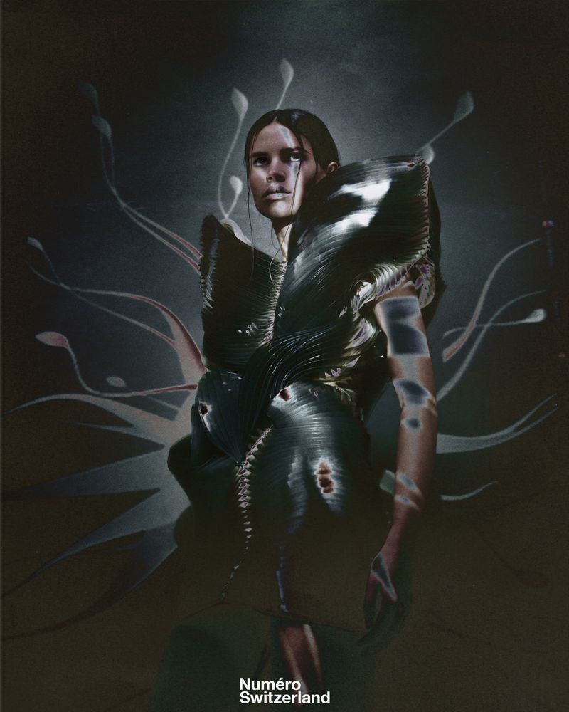 Fashion shot of a woman with dark hair wearing a sculptural, futuristic dark top against a dark background with light flourishes.