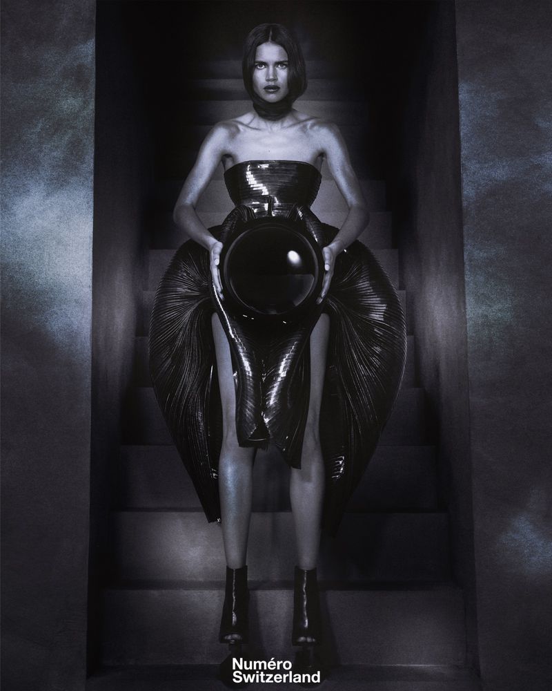 Fashionable woman poses on stairs wearing strapless dress and boots while holding a black orb in studio setting with muted color palette.