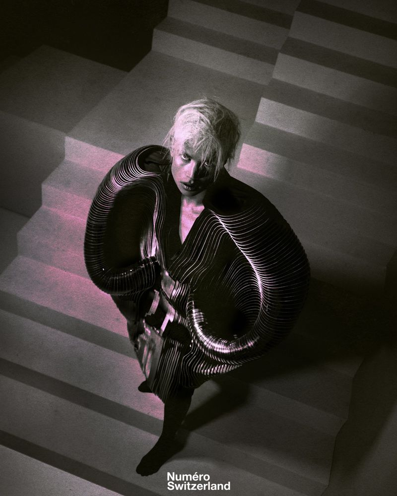 An adult female model with white hair poses on white stairs, wearing a futuristic black outfit with light-reflecting details in a fashion editorial.