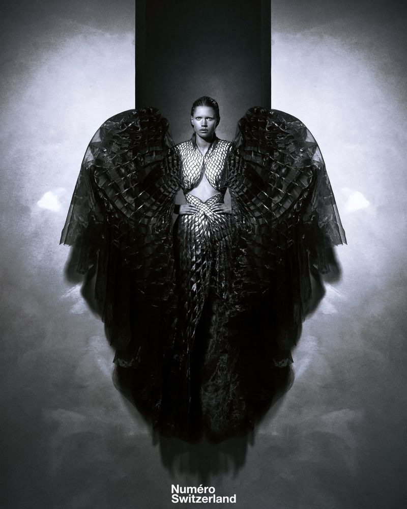 Monochrome fashion shot of a woman posing with wing-like costume element and textured dress against a smoky grey background.