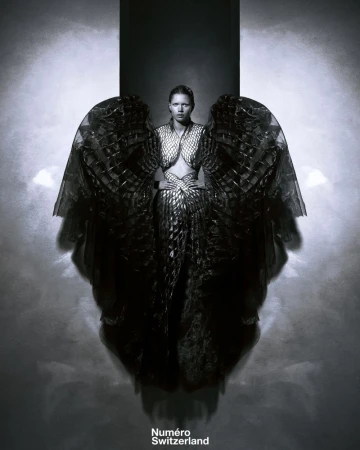 Monochrome fashion shot of a woman posing with wing-like costume element and textured dress against a smoky grey background.