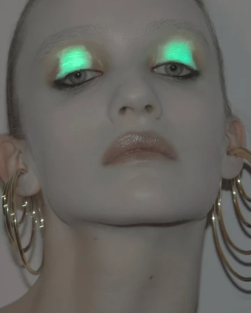 Close-up of a woman's face with glowing turquoise eye makeup and glossy lips, accented by large, tiered gold hoop earrings. Soft lighting.