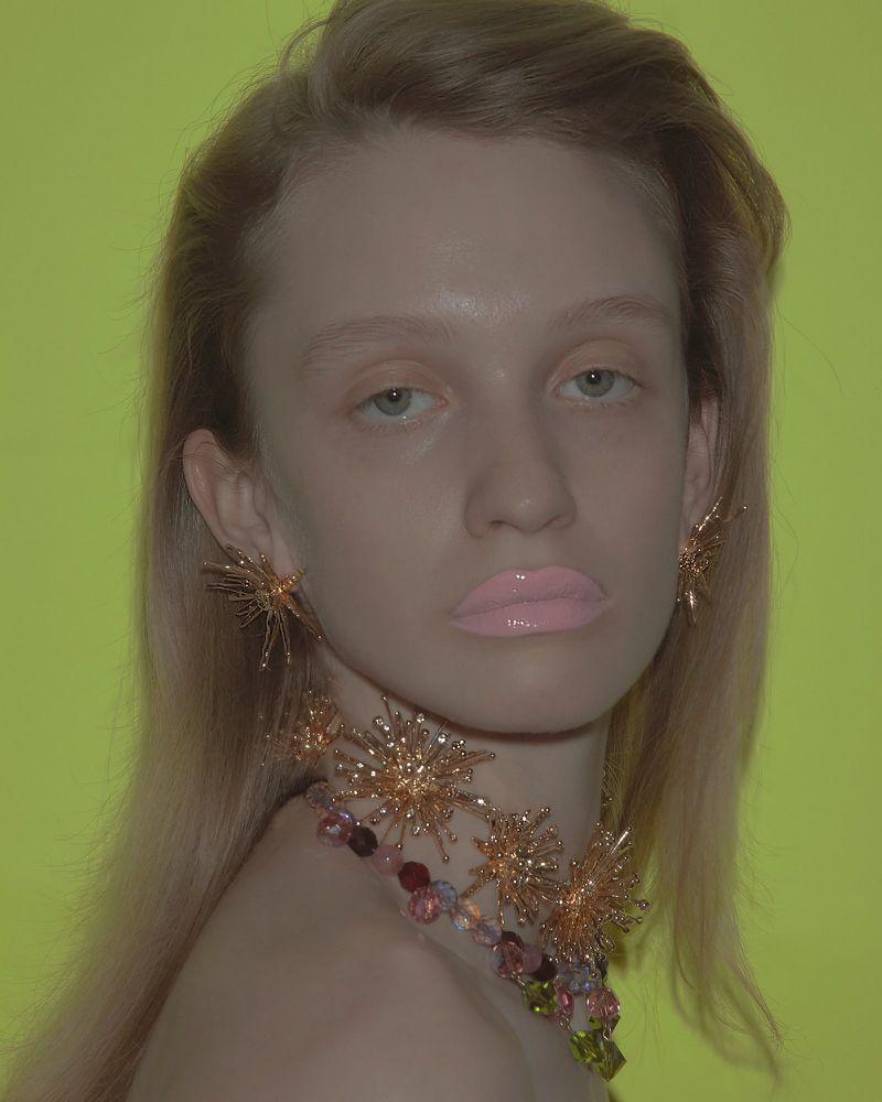 Woman with blond hair wearing a beaded choker and starburst earrings against a bright lime green background in a studio shot.