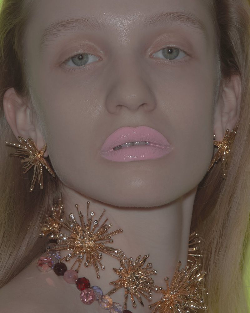 Close-up portrait of a fair-skinned young adult woman with striking golden starburst jewelry, glossy pink lips, and light eyes.