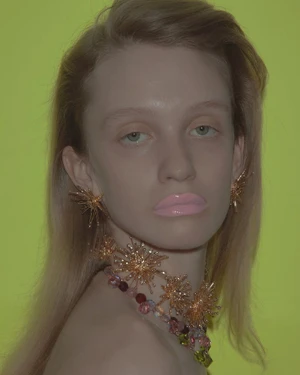 Close-up portrait of a model wearing glossy pink lipstick and ornate gold earrings against a neon yellow background.