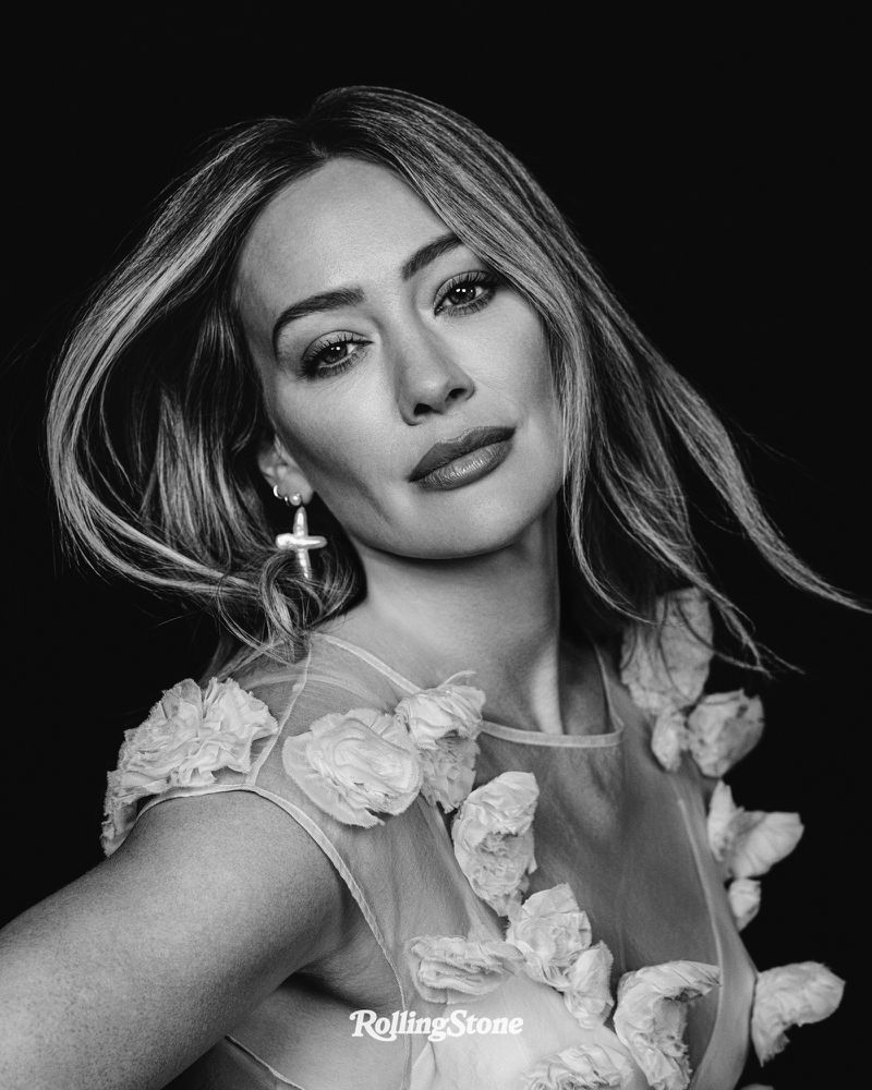 Black and white portrait of Hilary Duff in a floral dress against a dark background, showcasing her blonde hair and cross earrings.