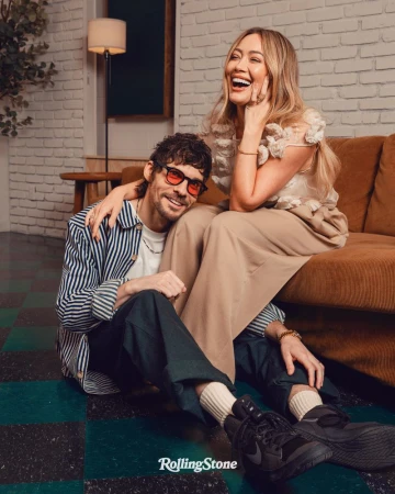 Candid photo of Hilary Duff sitting on a couch next to a man in glasses wearing a striped shirt while sitting on the floor.
