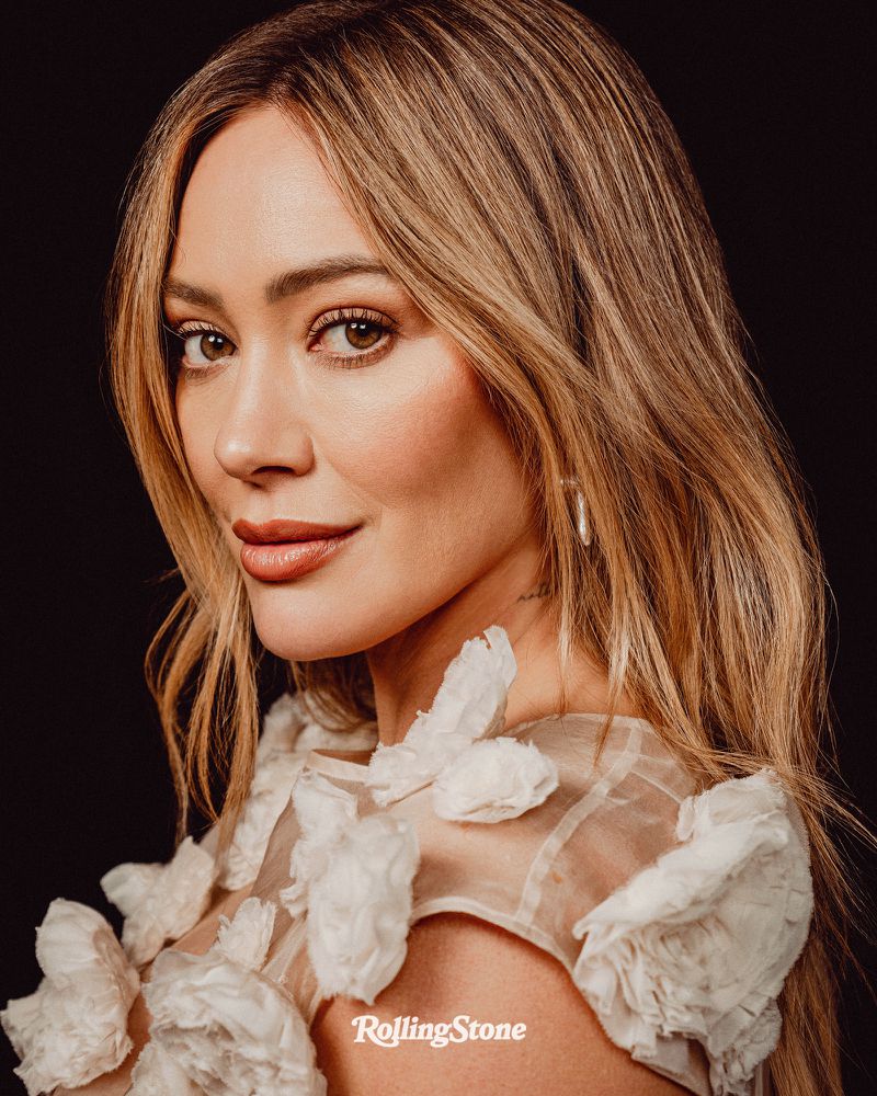 Close-up portrait of Hilary Duff with shoulder-length blonde hair, a flower-adorned top, and a dark background, showcasing a soft, elegant aesthetic.