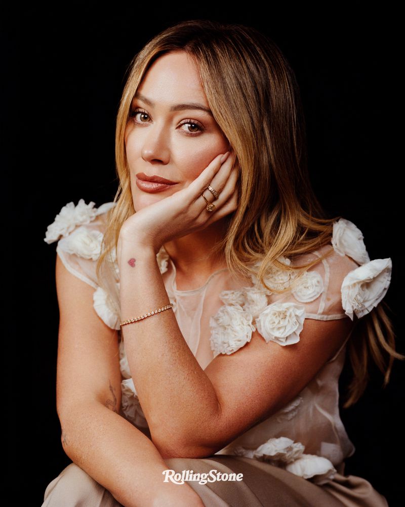 Portrait of Hilary Duff, gazing forward with hand to face, wearing a cream-colored dress with floral details against a black background.
