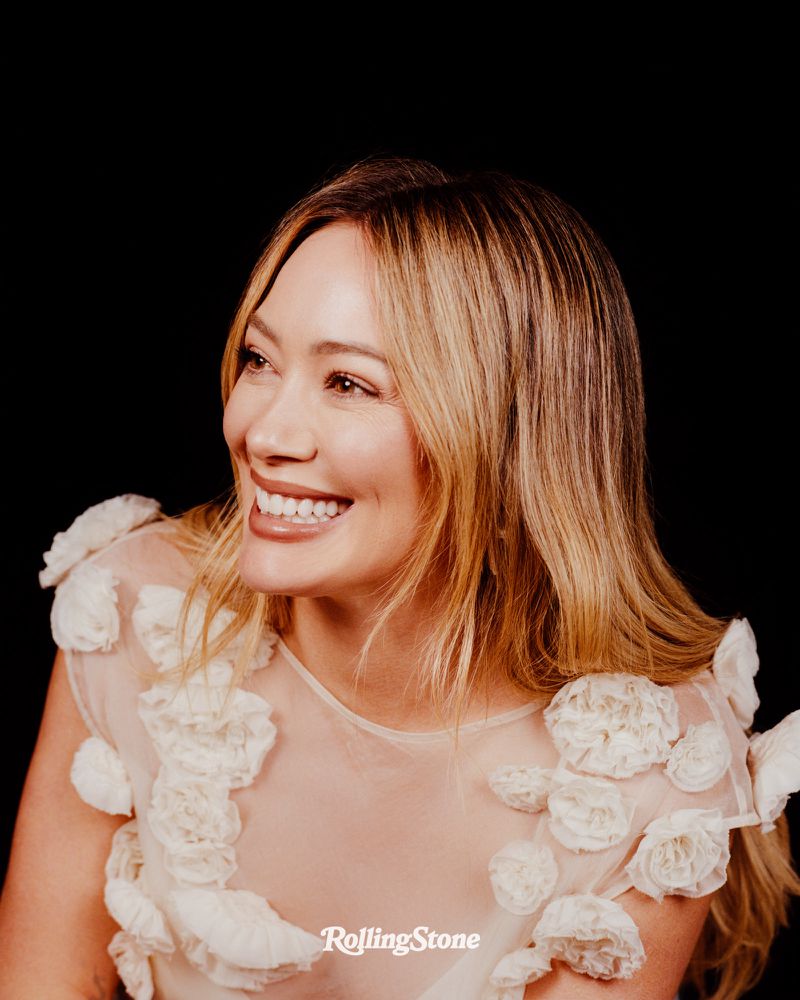 Close-up portrait of Hilary Duff smiling and looking to the side with a black background and wearing a sheer floral dress.