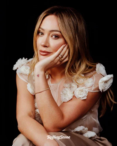 Hilary Duff posing in a cream floral appliqué top against a black background for Rolling Stone