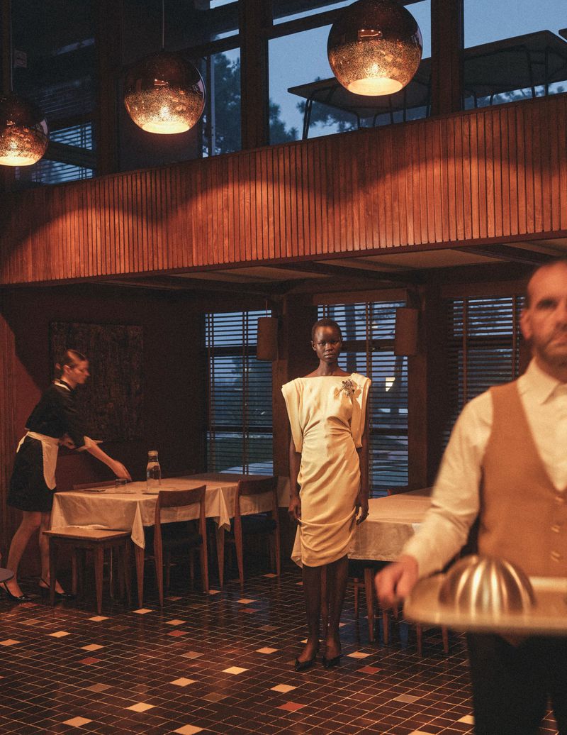 Elegant fashion shot in an upscale restaurant featuring an African model in a cream dress with a waiter and restaurant server.