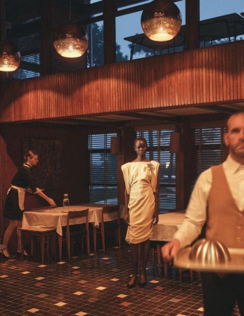 Elegant fashion shot in an upscale restaurant featuring an African model in a cream dress with a waiter and restaurant server.