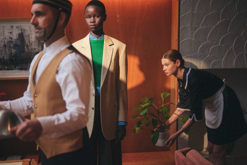 Indoor fashion shot showcases a woman in an oversized blazer with two hotel staff in the foreground and background.