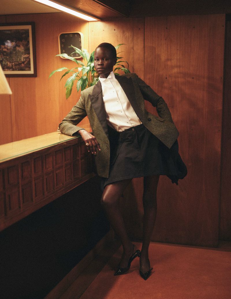 Woman poses indoors wearing a green blazer, white button-down shirt, and black skirt, complemented by black heels and a leafy green plant.