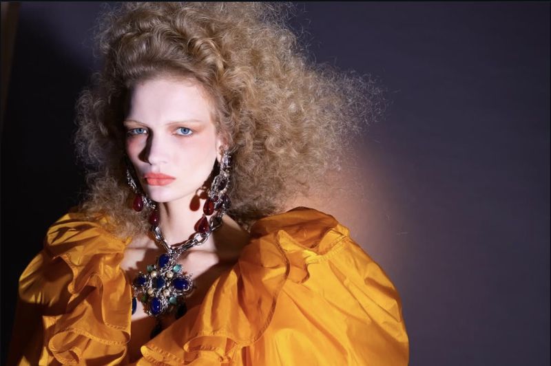 Blonde woman poses in yellow gown with statement jewelry in a studio shot against a dark backdrop with side lighting.