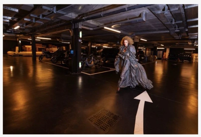 A woman with curly blonde hair in a flowing, ruched grey dress walks through a parking garage with arrow floor signage.