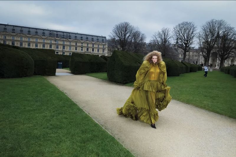 Woman in flowing olive gown walks along a stone path in green garden near historic building under cloudy sky in an outdoor fashion shoot.