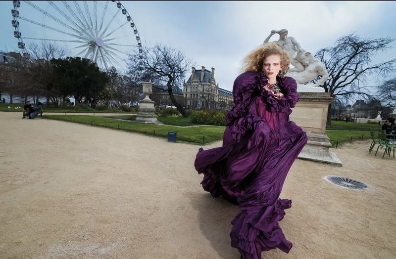 Fashion shot of a woman with blonde curly hair wearing a purple gown in an outdoor park setting with Ferris wheel in background.