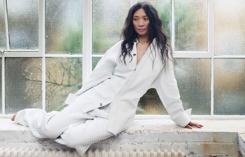 Adult woman with long dark hair in white textured suit sits on a windowsill in front of a frosted glass window with greenery outside.