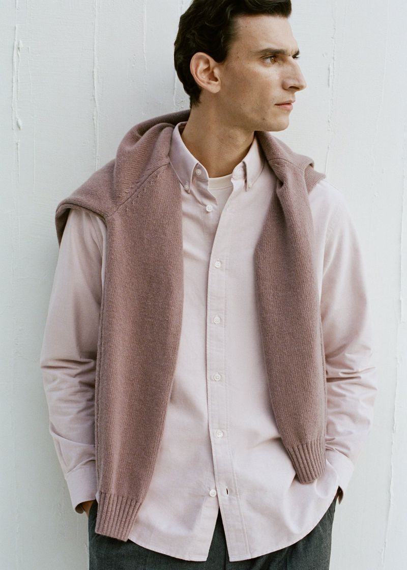 A fair-skinned adult male in a pink button-down shirt poses indoors against a white wall, wearing a mauve sweater around his shoulders.
