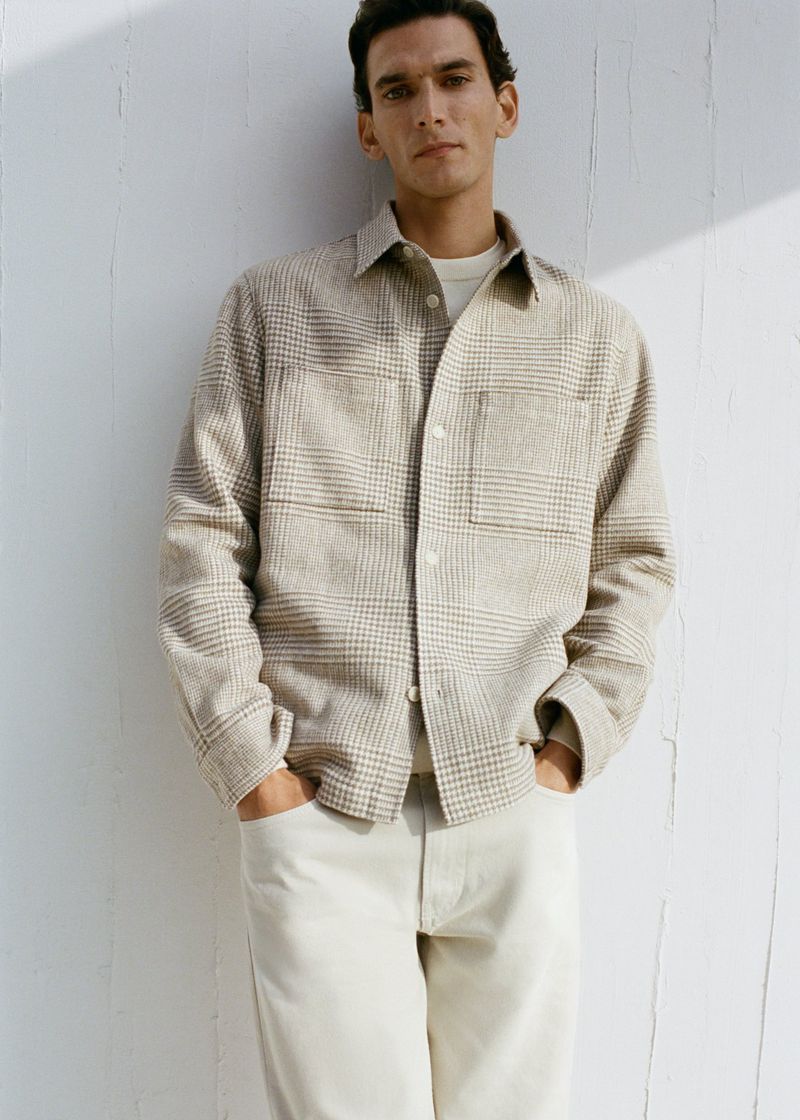 A man with hands in his pockets leans against a white wall, wearing a beige plaid shirt jacket, off-white t-shirt, and off-white trousers.