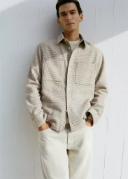 Man wearing a grey plaid overshirt and light trousers posing against a plain white wall in soft natural light.