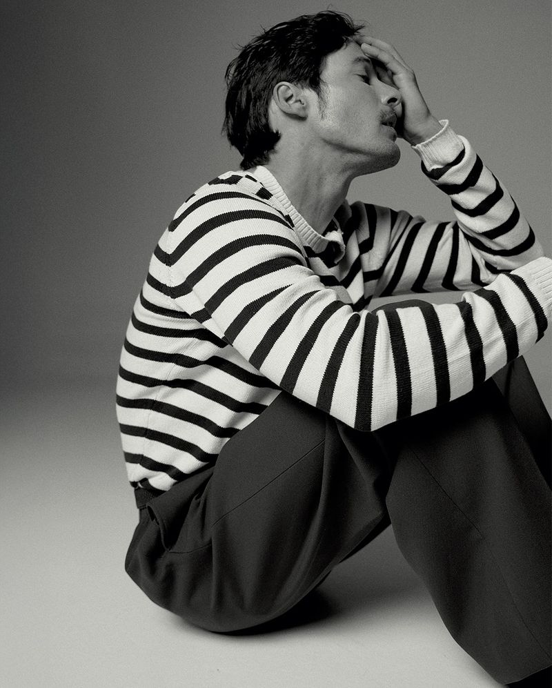 Portrait of a thoughtful adult male with hand on forehead, wearing striped sweater and dark pants, in a neutral studio setting.