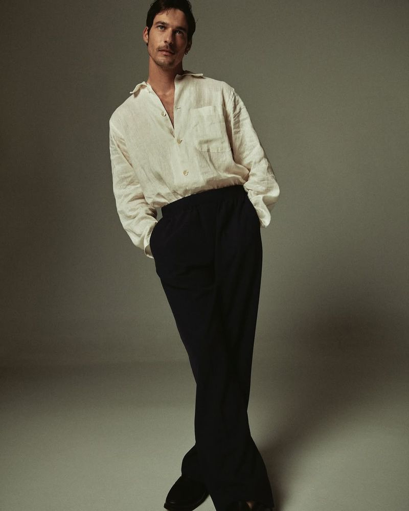 A handsome man stands confidently in a photo studio, posing in a casual white linen shirt and dark trousers, bathed in soft lighting.