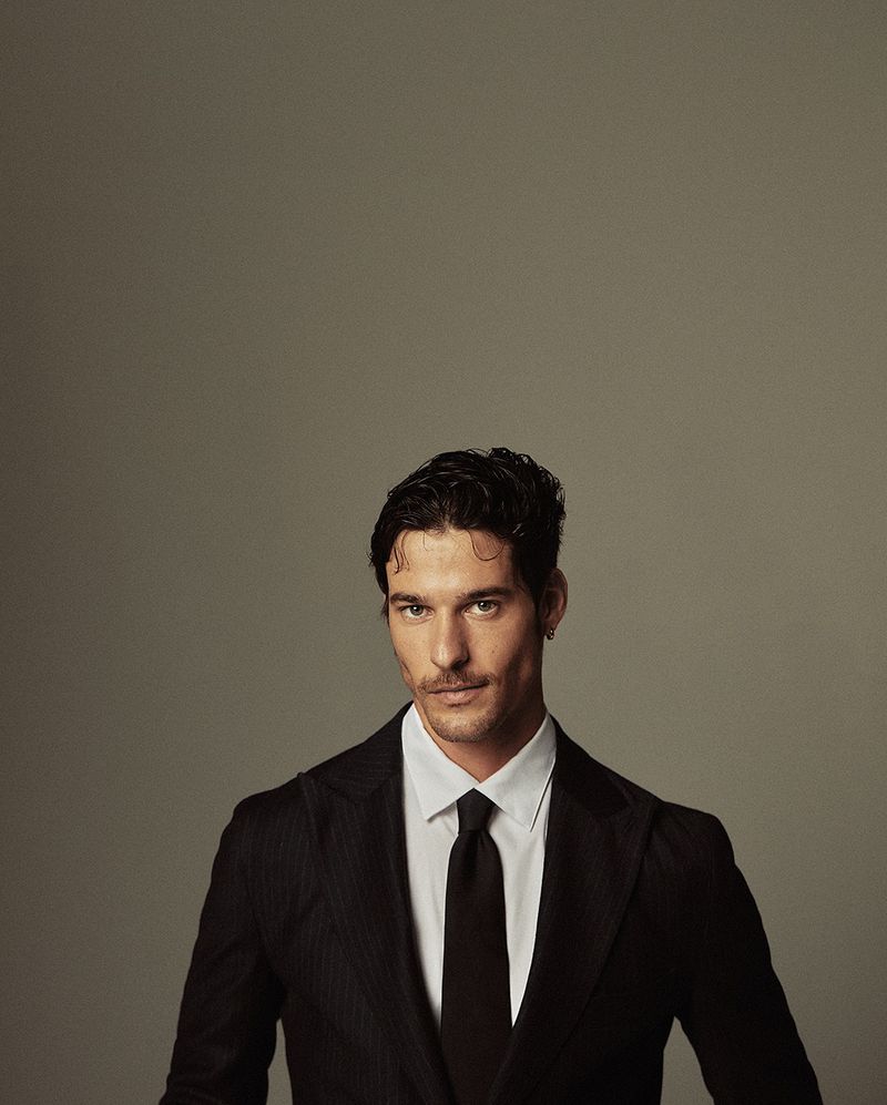 Portrait of a dark-haired adult man in a black suit and tie, posing against a neutral grey background in a studio setting. He is centered.
