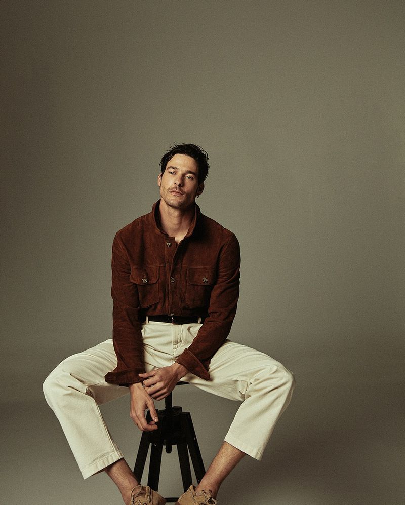 Portrait of a man seated on a stool, wearing a brown corduroy shirt and beige trousers in a neutral studio setting.