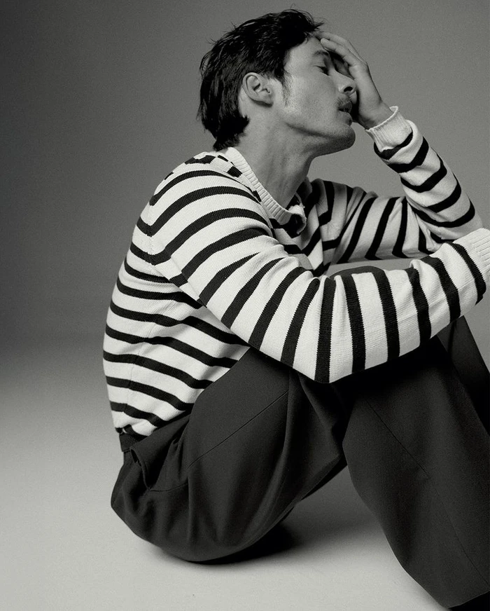 Man wearing a striped sweater and dark trousers sitting on the floor with hand to forehead in a studio.