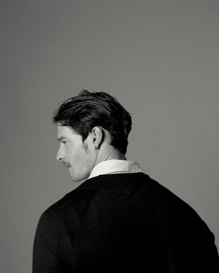 Profile portrait of a man with dark hair and a mustache wearing a black jacket and white collared shirt against a grey background.