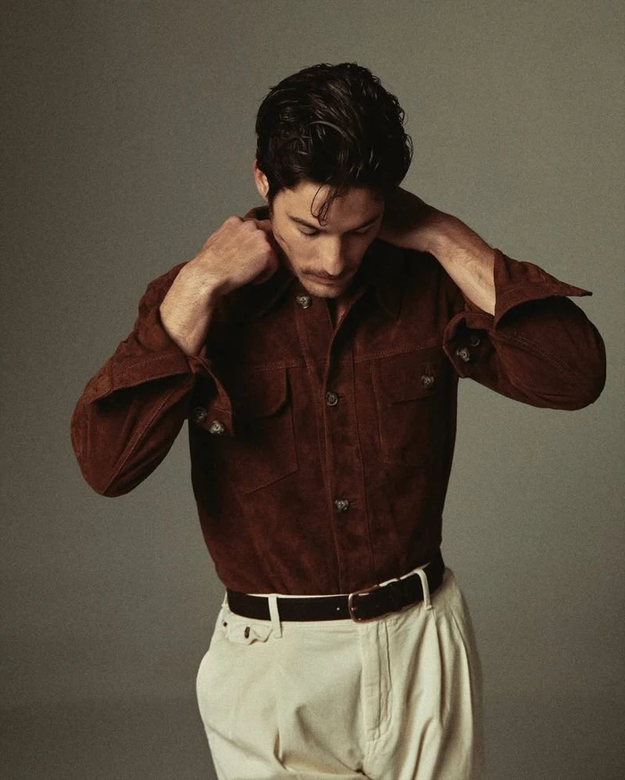 Man adjusting his brown suede shirt wearing white pleated trousers against a neutral grey studio background
