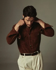 Man adjusting his brown suede shirt wearing white pleated trousers against a neutral grey studio background