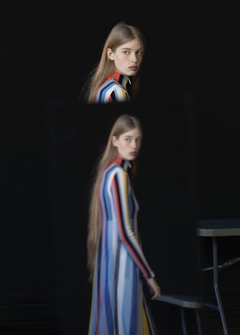 Woman poses in a striped dress in a studio with a black backdrop and a stool, captured in a fashion photography style with soft lighting.