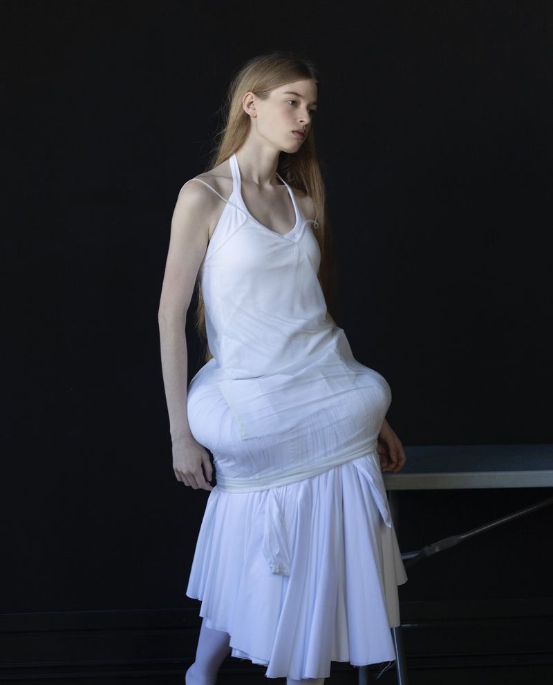 A solitary woman wearing a white dress poses in front of a dark background next to a metal table; indoor fashion studio photograph.