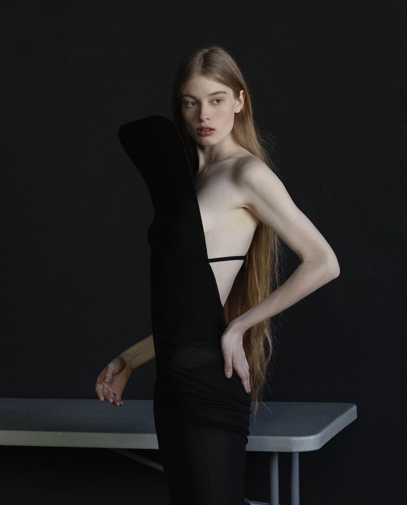 A slender woman poses in a black dress with dramatic shoulder detail in a studio with a gray table and dark background.