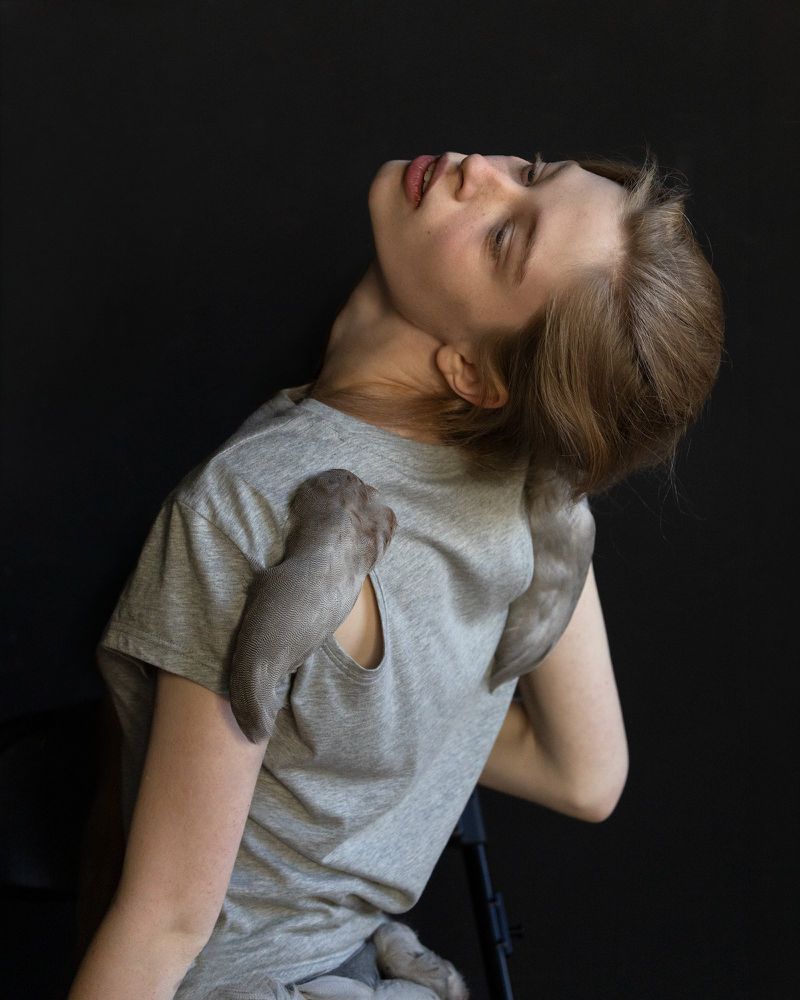 Young woman tilts head back in a studio, wearing grey shirt, two puppies nestled on her shoulders, black background, soft lighting.