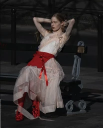 Young woman in a layered sheer pink dress with a red sash and red shoes seated on a dark park bench outdoors