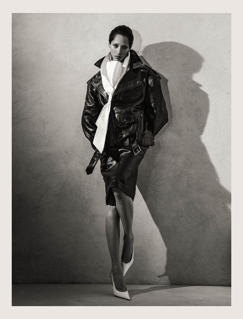Elegant woman in a leather jacket, skirt, and white heels poses against a textured wall with soft lighting, creating a dramatic fashion portrait.
