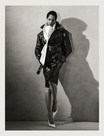 Elegant woman in a leather jacket, skirt, and white heels poses against a textured wall with soft lighting, creating a dramatic fashion portrait.