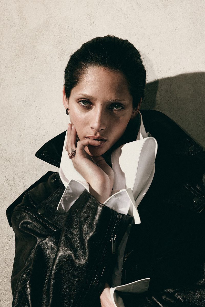 Close-up portrait of an adult female with her hand touching her face. She is wearing a black leather jacket and a white ruffled shirt.