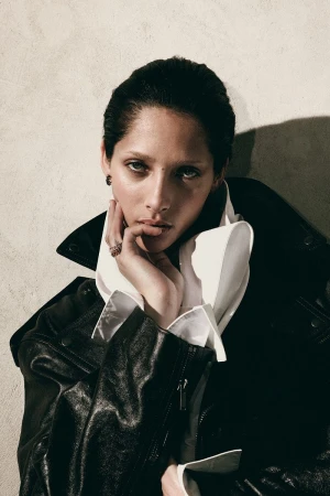 Close-up portrait of an adult female with her hand touching her face. She is wearing a black leather jacket and a white ruffled shirt.