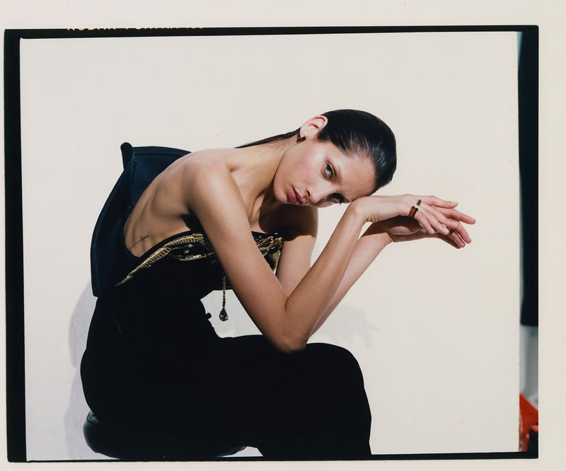Elegant brunette woman with her arms on the back of a chair, wearing black dress with gold detail, shot in a photo studio.